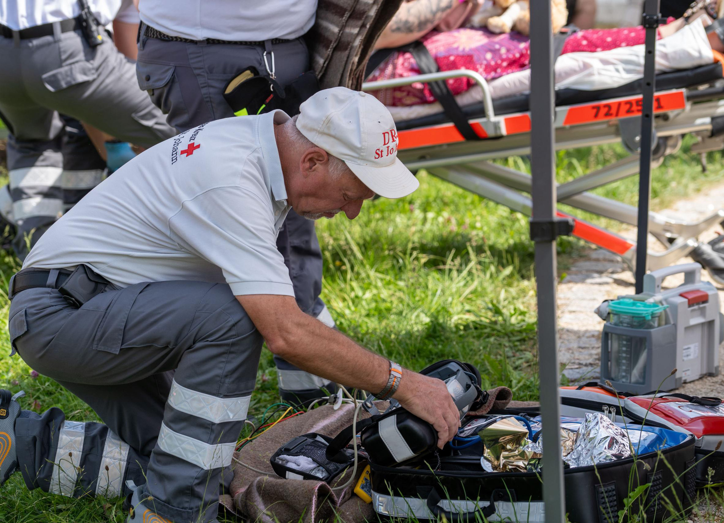 Auf dem Bild ist ein Helfer knieend vor einem Sanitätsrucksack zu sehen. bei der Gruppenarbeit im Freien zu sehen.  im Garteniche Helferinnen und Helfer aus den Bereitschaften Eningen und Pfullingen sowie Fahrzeuge des Bevölkerung- und Katastrophenschutzes zu sehen.